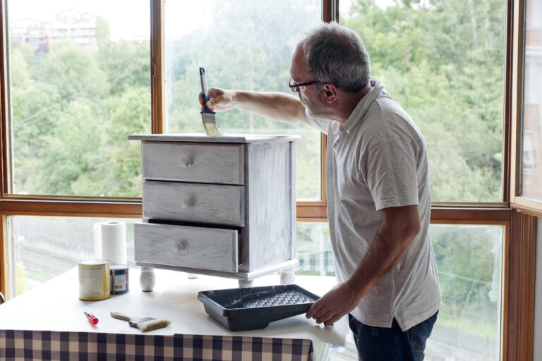 Un hombre mayor con gafas pintando un mueble con pintura blanca.